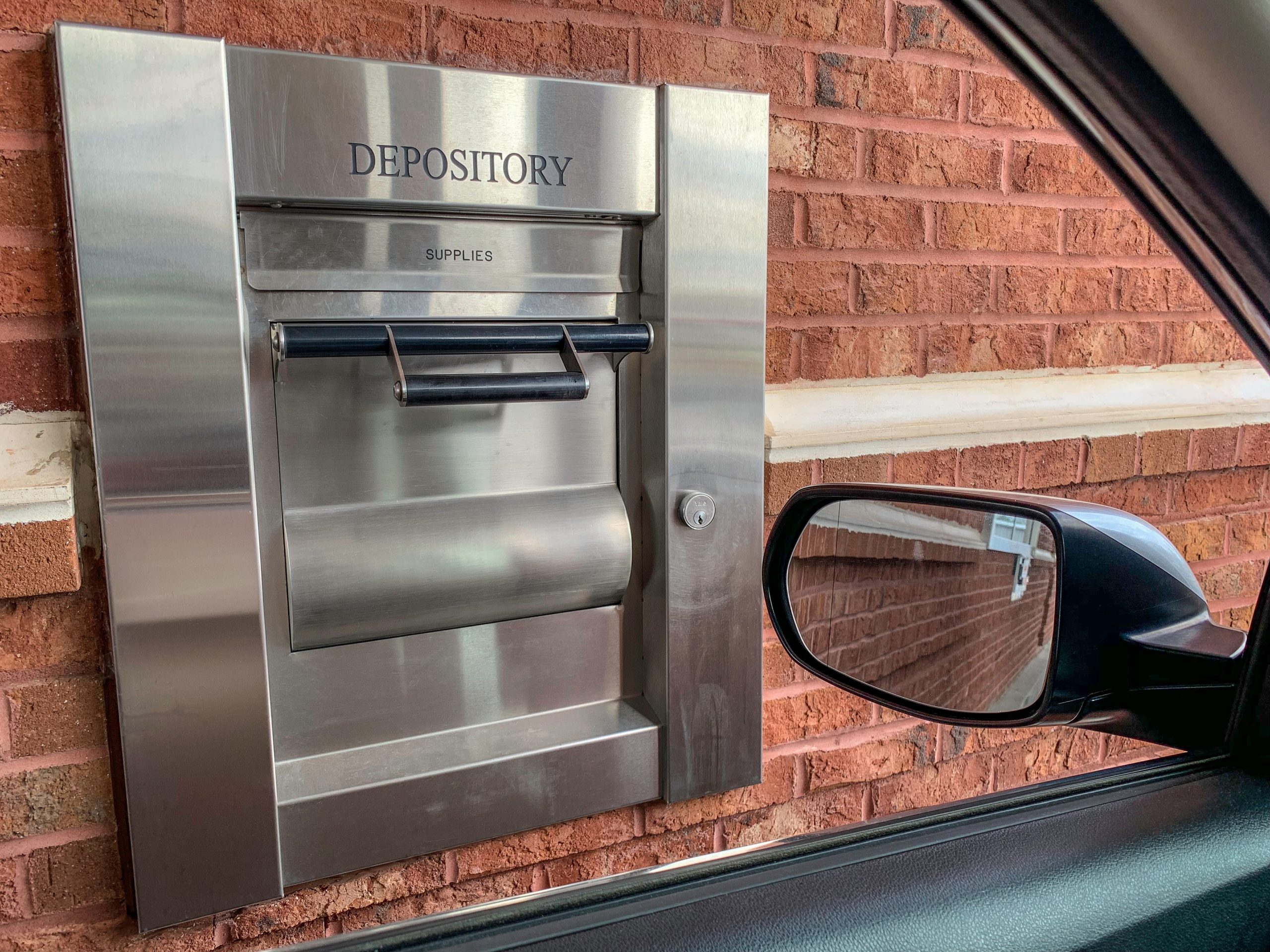 The 24-hour drive-thru night depository of a neighborhood bank set on a brick wall and seen through the car window in a horizontal image format.